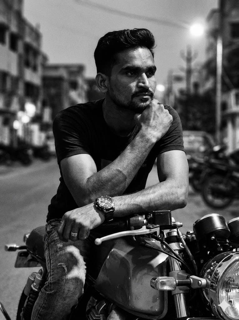 Nano Banana generated image: A black-and-white portrait with a rugged, cinematic aesthetic captures a male figure seated on a classic motorcycle (café racer or bobber style). The subject is leaning forward over the handlebars, striking a thoughtful and intense pose. One hand rests on the headlight or handlebars, while the other arm is bent, supporting his head. His gaze is directed outward, conveying a contemplative and virile expression.
He wears a dark cotton T-shirt and pants (possibly jeans). A chunky wristwatch is visible on one arm, and a ring on his finger.
His hair is medium-length, textured, and casually tousled. A short, well-groomed beard defines his face.
The background is a softly blurred urban or street environment. The lighting is natural and dramatic, with high contrast, creating deep shadows and sharp highlights that sculpt the facial features and arm muscles. The image has a subtle grain, evoking the look of analog film.
Camera Settings: Captured with a prime portrait lens (e.g., 85mm or 105mm) on a full-frame camer